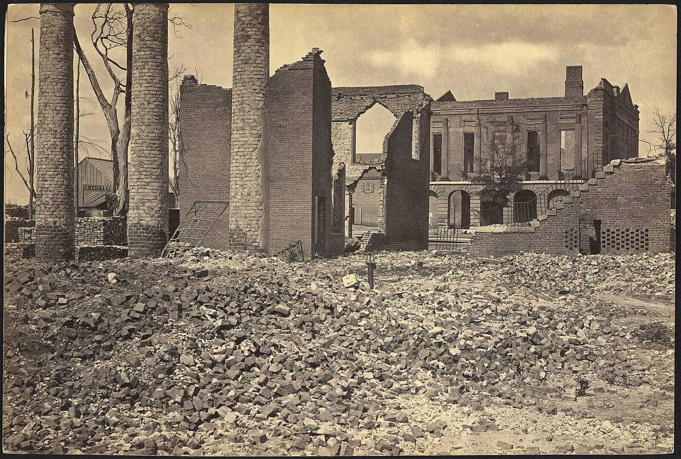 Photograph of a burned city block in 1865: brick chimney stacks and roofless walls standing in a field of rubble, with the shell of a large colonnaded building behind