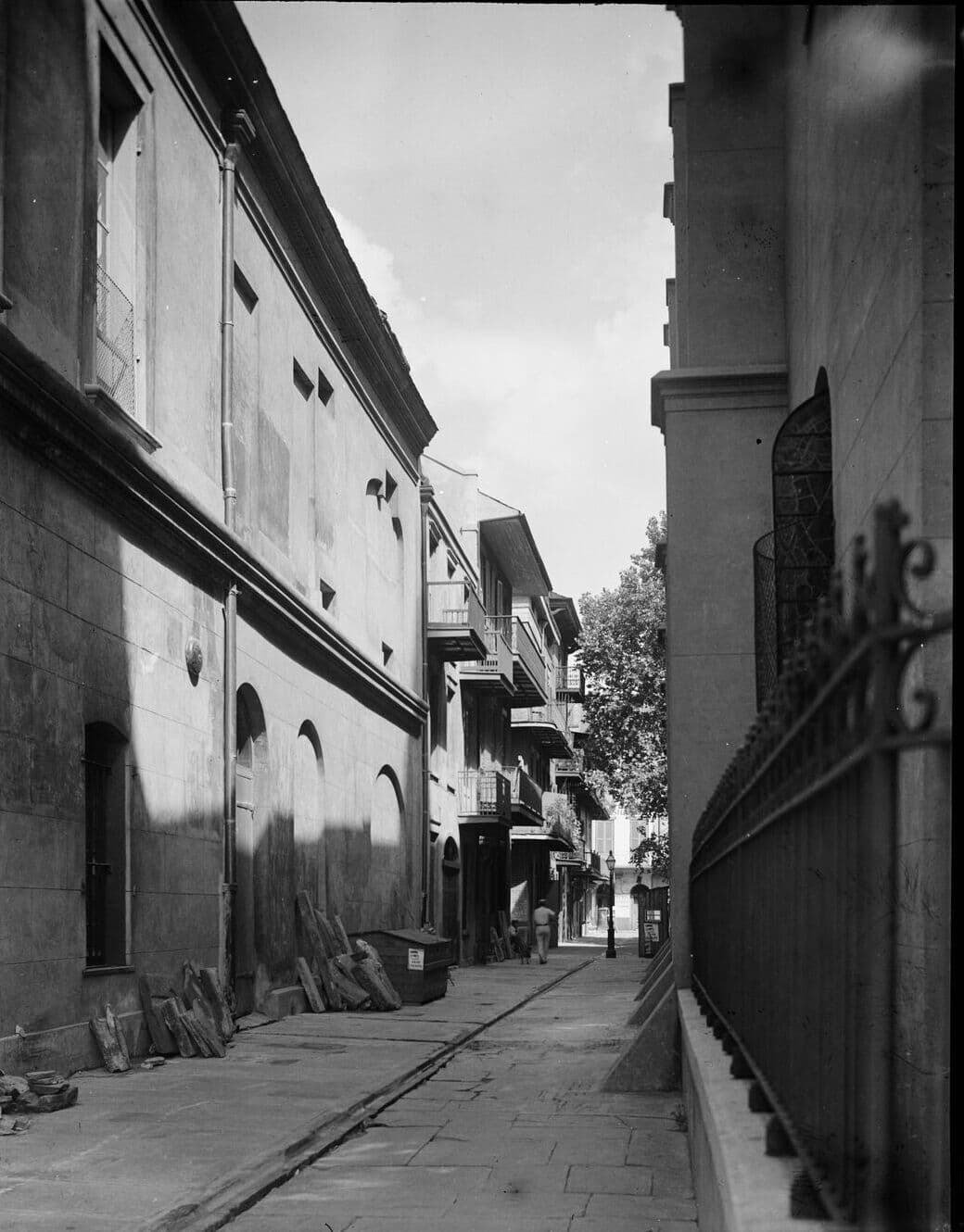 Black-and-white photograph looking down a narrow flagstone alley beside the Cabildo in New Orleans, its plastered wall and arched windows on the left, iron railings on the right, and balconied houses at the far end