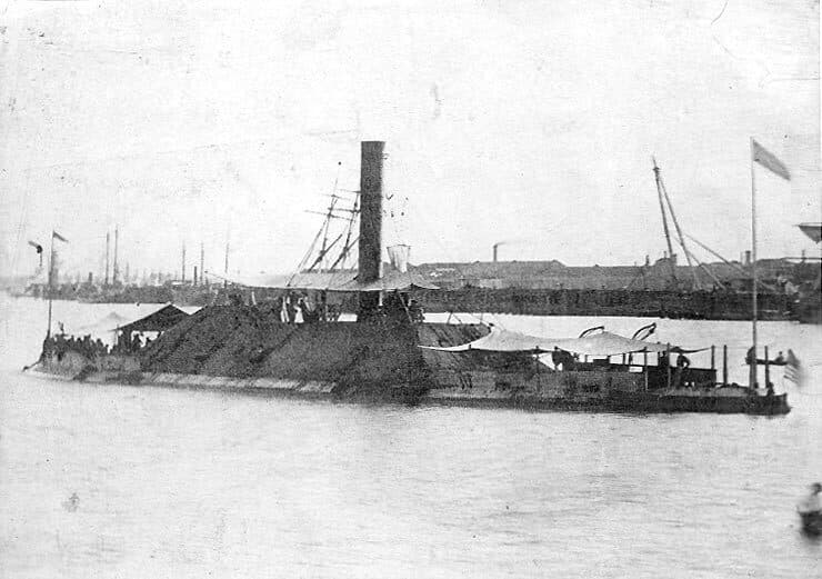 Faded 1865 photograph of the ironclad Tennessee at anchor: a long, low hull with a sloped armoured casemate, a single tall funnel and awnings rigged over the deck, with a wharf and buildings behind