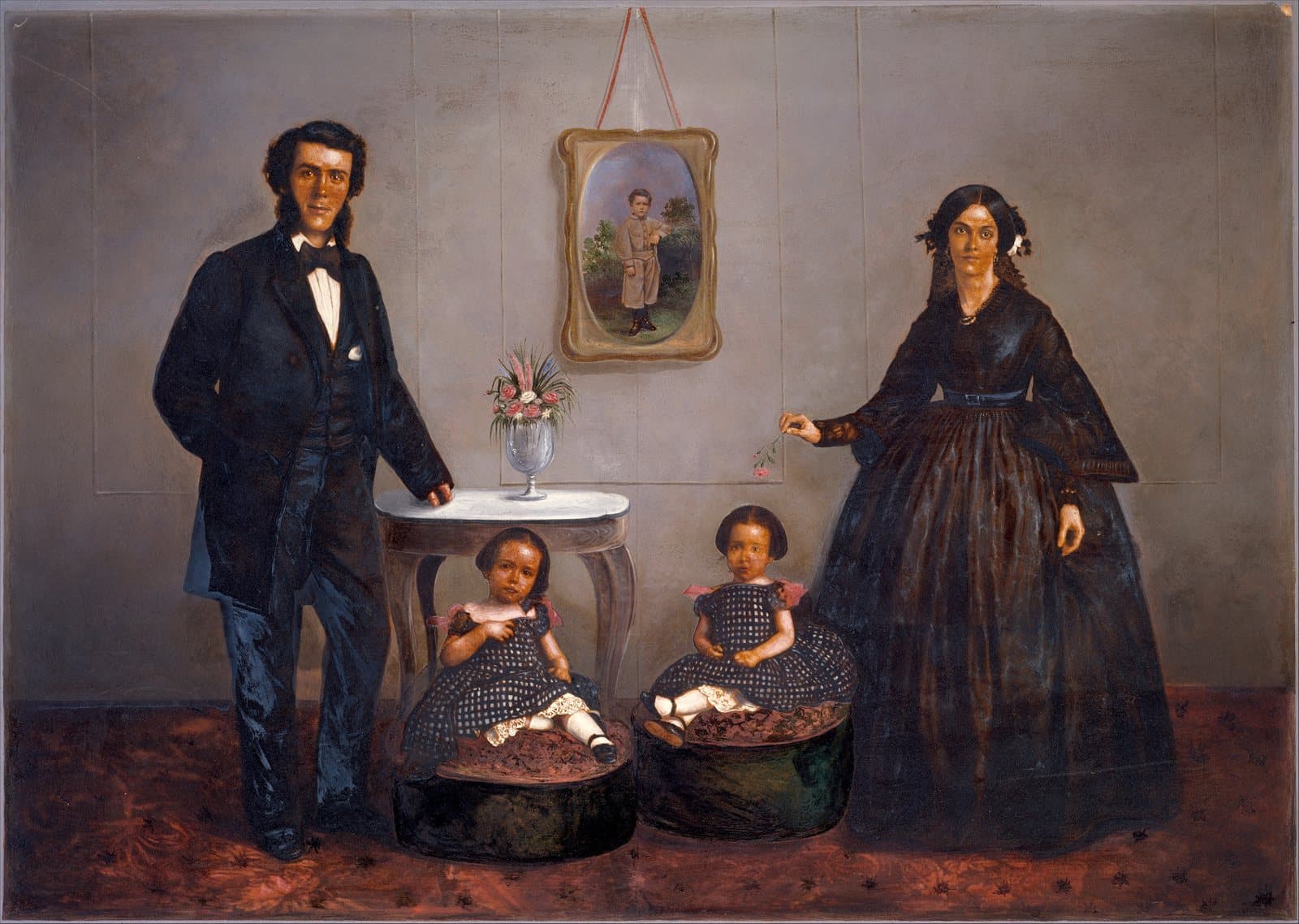 Hand-coloured 1850s photograph of a family in a parlour: a father in a dark suit, a mother in a black dress, and two small daughters in matching checked dresses seated between them, with a framed portrait of a boy on the wall