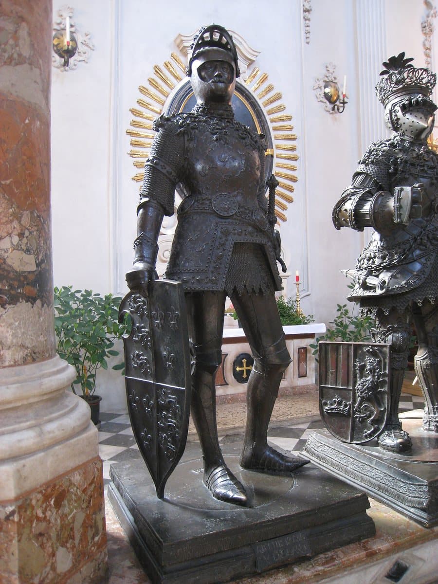 Life-size bronze statue of a knight in armour holding a shield, standing on a stone plinth inside a church, beside another armoured bronze figure