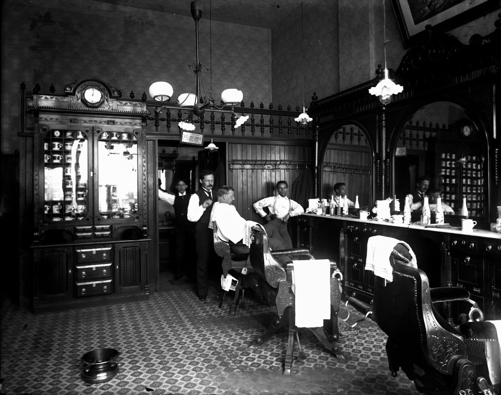 Black-and-white photograph of a hotel barber shop in the 1890s: a barber in a waistcoat shaving a customer draped in white, ornate mirrored cabinets of shaving mugs, and a row of leather barber chairs on a tiled floor