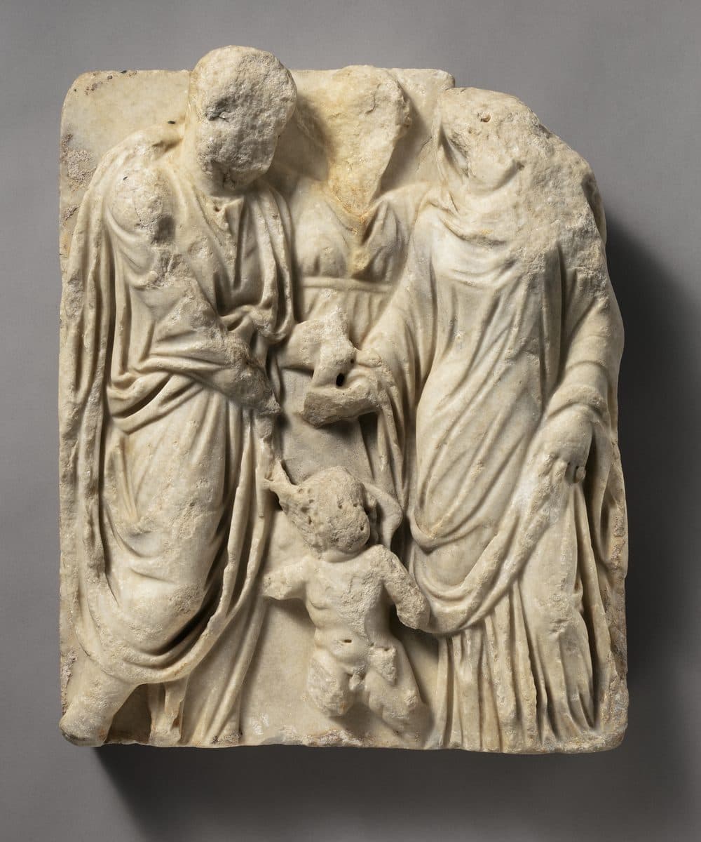 Weathered marble relief of a Roman man in a toga and a veiled woman joining right hands, with a figure standing between them and a small winged child at their feet
