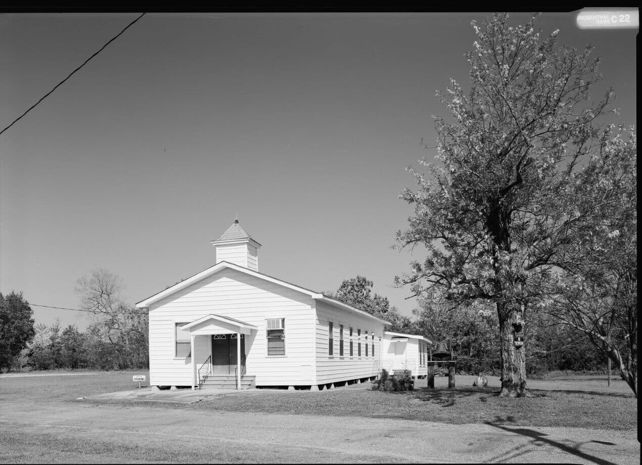 Black-and-white photograph of a small white clapboard Baptist church with a square belfry, a bell on a post beside it and a large tree in the yard